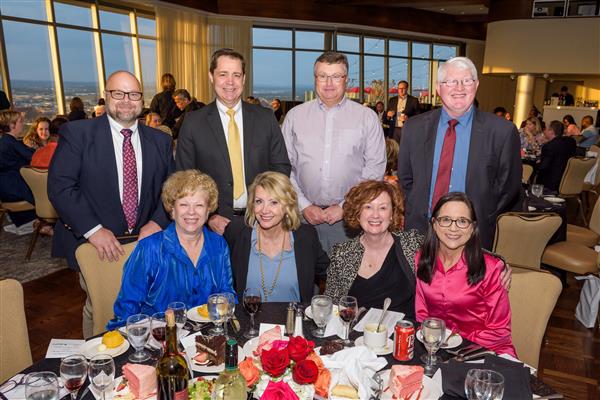 A group of eight well-dressed adults pose for a photo at a formal dinner event. Four individuals are standing behind a round table while four others are seated at the table. The table is set with glasses, plates of cake, salads, rolls, and bottles of wine and water. The background shows a large room